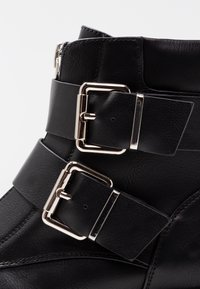 Bota de cuero negra con dos hebillas y correas de metal plateado, cremallera visible en la parte trasera, costuras detalladas en la superficie de cuero liso.