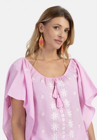 Top rosa ricamato con maniche larghe, caratterizzato da motivi floreali e frange sullo scollo. Abbinato a orecchini di perline multicolori.