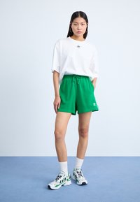 Hvit kortermet t-skjorte med liten svart logo, sammen med grønne shorts og hvite joggesko med grønne detaljer. Flate, glatte teksturer.