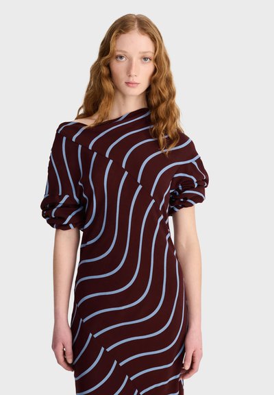 Jeune femme aux longs cheveux roux porte une robe violet foncé avec des rayures ondulées bleu clair, une épaule découverte et des manches froncées.