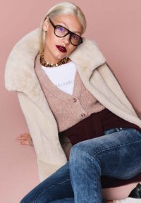 Strickpullover in pinken Tönen, kombiniert mit einem cremefarbenen Kunstpelzmantel. Dunkle Jeans und eine auffällige, kräftige goldene Kette als Akzent. Schwarze Brille und kräftige Lippen.