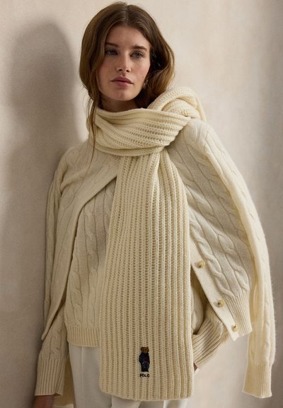 Kremasto-bež pleten šal z rebrasto teksturo in logotipom medveda. Združeno s puloverjem z vzorcem kabelske pletenine, ki izžareva prijetno zimsko zasnovo.