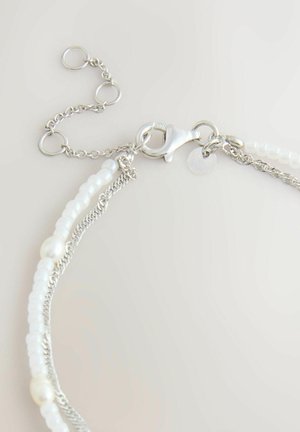 Srebrna bransoletka z łańcuszkiem, białymi koralikami i perłami, z zapięciem typu karabińczyk i regulowanym pierścieniem na jasnym tle.