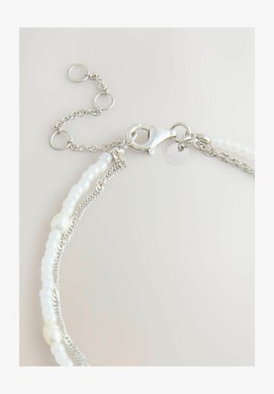 Srebrna bransoletka z łańcuszkiem, białymi koralikami i perłami, z zapięciem typu karabińczyk i regulowanym pierścieniem na jasnym tle.