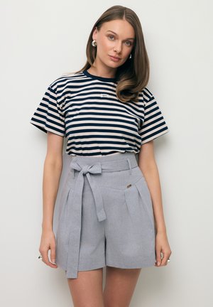 Kobieta o prostych brązowych włosach, nosząca srebrne kolczyki-koła, granatowo-białą koszulkę w paski oraz jasnoszare szorty z wysokim stanem i materiałowym paskiem z kokardą.