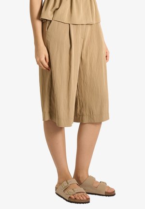 Pantaloni beige a gamba larga di lunghezza media indossati con sandali beige con cinturino e fibbia su un modello in piedi davanti a uno sfondo bianco.