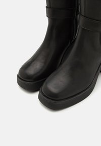 Bottes en cuir noir avec une finition lisse, bout rond, semelle à plateforme basse et fermeture à glissière sur le côté. Présentent une sangle ajustable au niveau de la cheville.