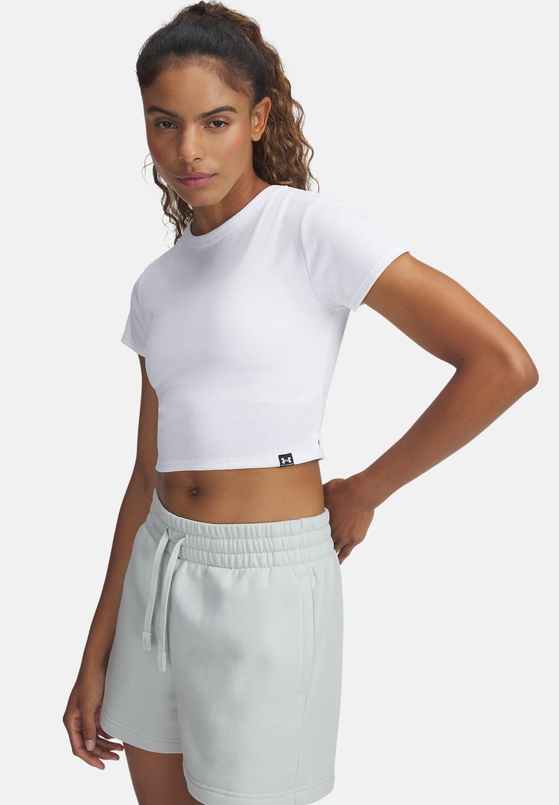 Kvinna med lockigt hår som bär en vit kortärmad t-shirt och ljusgrå shorts med dragsnöre, stående med en hand på höften.