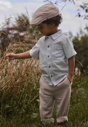 Jauns bērns ar bēša krāsas cepuri un gaismas kreklu maigi pieskaras garai zālei dabiskā āra vidē.