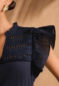 Top navy cu smocking texturat pe corp, mâneci transparente cu volane și gât înalt, evidențiind detaliile intricate și un finisaj neted.