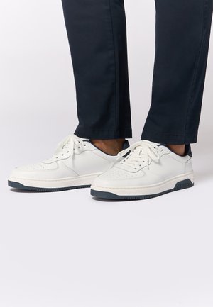 Weiße Leder-Sneaker mit Perforationen, Schnürung vorne und marineblauen Akzenten. Sie verfügen über eine dicke weiße Sohle mit einem strukturierten schwarzen Boden.