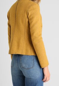 Femme portant une veste cintrée jaune moutarde et un jean en denim bleu, vue de dos et de côté sur un fond uni.