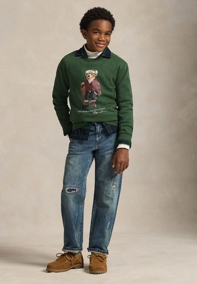 Grön sweatshirt med björngrafik, blå rutig krage, slitna jeans av denim och ljusbruna moccasinliknande skor. Enkel bakgrund.