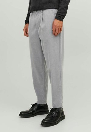 Mann trägt hellgraue, plissierte Hose, schwarze Schnürschuhe, dunkles, strukturiertes Langarmshirt und steht vor einfarbigem Hintergrund.