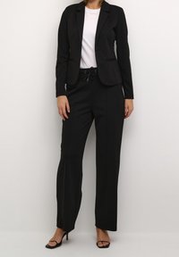 Schwarzer taillierter Blazer mit Reverskragen, kombiniert mit lockeren schwarzen Hosen mit Kordelzugbund. Stylisch mit schwarzen High Heels.