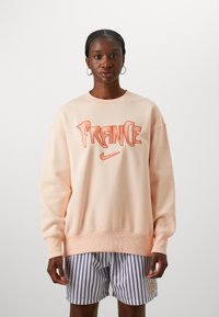 Sudadera melocotón con la palabra "FRANCE" en rojo y el logo de Nike, combinada con pantalones cortos a rayas. Material de algodón con puños y dobladillo acanalados.