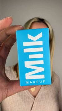 Hand holding a blue Milk Makeup box with white text, partially covering a person wearing a beige top and a gold necklace.