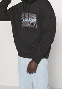 Felpa nera con cappuccio realizzata in tessuto morbido, dotata di una tasca frontale e un motivo grafico a linea ondulata bianca e blu con il testo "Calvin Klein".