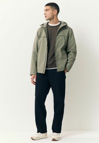 Bărbat îmbrăcat cu o jachetă cu glugă de culoare verde deschis, un pulover închis la culoare, pantaloni negri și teniși albi, stând cu o mână în buzunar, privindu-se în lateral.