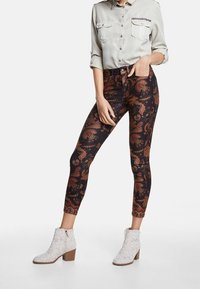 Leggings con estampado sobre un fondo oscuro que presentan vibrantes diseños de paisley en rojo, naranja y verde; combinados con una camisa clara de botones y botas blancas hasta el tobillo.