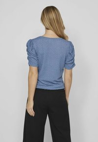 Modrá textúrovaná blúzka s krátkymi pufovanými rukávmi a okrúhlym výstrihom, skombinovaná s čiernymi nohavicami s vysokým pásom. Hladká tkanina a priliehavý strih.