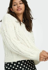 Pullover bianco a maglia con motivo a treccia e vestibilità ampia, con polsini a coste, abbinato a una gonna nera con pois. Texture morbida, design informale.