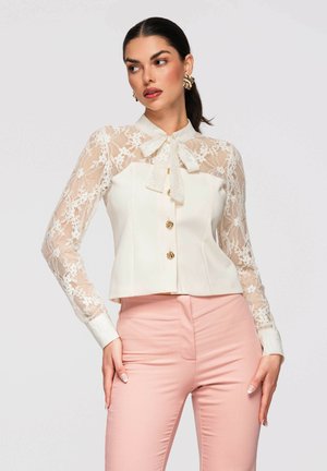 Kobieta w białej koronkowej bluzce z długim rękawem, zdobionej złotymi guzikami i kołnierzykiem z kokardą, w spodniach z wysokim stanem w jasnoróżowym kolorze, pozująca z rękami na biodrach.
