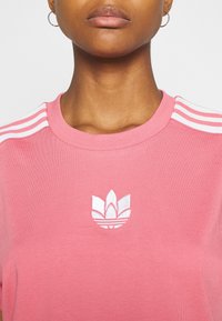 Rosa bomullströja med rund halsringning, vita tre-stripes accenter på axlarna och en broderad vit logotyp på bröstet.