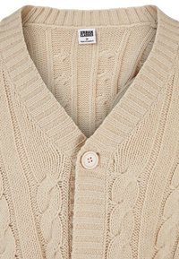 Cárdigan de punto beige con escote en V, que presenta patrones de trenzas texturizadas, bordes acanalados y un cierre de un solo botón. Etiqueta: Urban Classics.