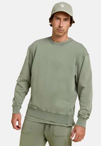 Sweatshirt vert olive en coton doux, avec des poignets et un ourlet côtelés, présentant un col rond et un petit logo sur le devant.