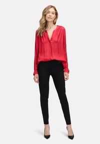 Blouse en soie rouge avec un décolleté en V profond, des manches longues et des poignets boutonnés. Associée à un pantalon noir ajusté et des talons hauts noirs.