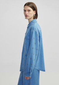 Camicia di jeans azzurro chiaro con chiusura a bottoni, maniche lunghe e dettagli di ricamo floreale sparsi su tutto il tessuto.
