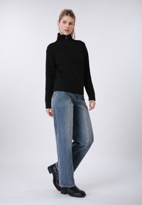 Pull noir en maille avec un col montant zippé, associé à un jean bleu clair à jambes larges et des bottines, illustrant un look décontracté.