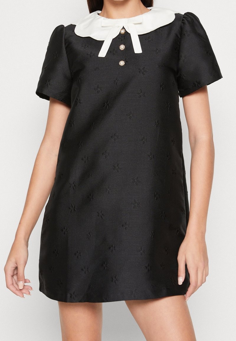Robe noire à manches courtes avec une texture florale subtile, col Peter Pan blanc, trois boutons en perle et un petit nœud décoratif blanc.