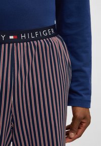 Σκούρα μπλε παντελόνια πιτζάμας με κόκκινες και λευκές κάθετες ρίγες, με σκούρα μπλε μέση που φέρει το λογότυπο "Tommy Hilfiger". Ορατό μακρυμάνικο μπλε πουκάμισο.