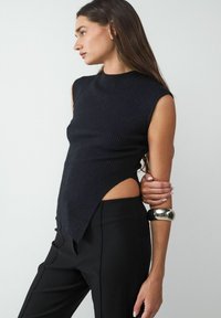 Schwarzes, geripptes, ärmelloses Oberteil mit hohem Kragen und asymmetrischem Saum, kombiniert mit schwarzen Hosen. Inklusive einer vergoldeten Armreifen.