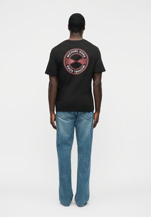 Férfi fekete Michael Kors pólóban, amelyen kör alakú logó van a hátulján, és kék farmert visel, egyszínű világos háttér előtt áll.