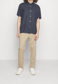 Modrá lněná košile s krátkým rukávem, límcem na knoflíky a náprsní kapsou, kombinovaná s béžovými bavlněnými kalhotami a bílými teniskami. Lehká textura.