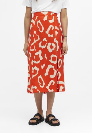 Femme portant une jupe midi taille haute orange avec un motif abstrait beige, un haut blanc, des sandales noires, debout sur fond blanc.