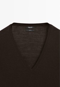 Pull en laine marron foncé avec un col en V. Présente une texture douce et des étiquettes minimalistes indiquant la marque et les informations sur le matériau.