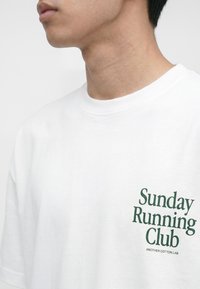 Primo piano di una persona che indossa una maglietta bianca con scritta verde "Sunday Running Club" e testo più piccolo "Another Cotton Lab" sul petto.