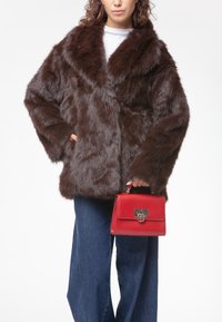 Manteau en fausse fourrure marron avec larges revers et manches longues, associé à un petit sac à main rouge avec fermeture à fermoir et accents argentés.