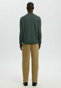 Persona de pie de espaldas con una camiseta de manga larga verde oscuro, pantalones de pana beige y zapatos negros, frente a un fondo sencillo.