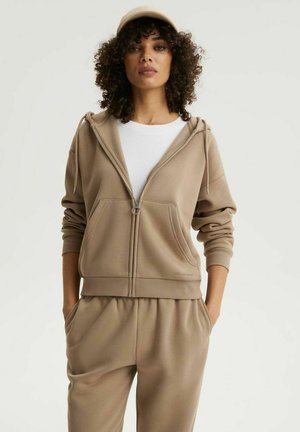 Kobieta o kręconych włosach, ubrana w beżową bluzę na suwak, dopasowane spodnie, beżową czapkę i białą koszulkę, stojąca z rękami w kieszeniach.