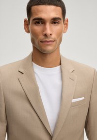 Junger Mann mit kurzen Haaren, der einen beigen Blazer über einem weißen Rundhalsausschnitt trägt, mit einem weißen Einstecktuch in der Brusttasche des Blazers.