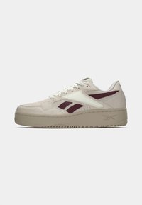 Zemi zemi beiges kedas ar baltām un tumši sarkaniem akcentiem, baltām saites un biezām beigtām zolei ar Reebok logotipu sānos.