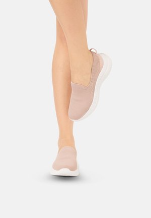 Benen met lichtroze gebreide slip-on sneakers met witte zolen, één voet opgeheven die de zijkant van de schoen en de zooldetails toont.