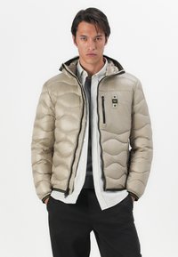 Beige puffer jacket with quilted pattern, black zipper, front pocket, and adjustable hood. Worn over a gray shirt and black pants.