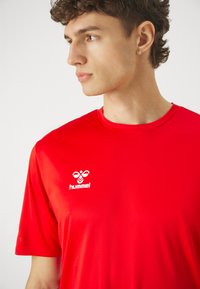 Rød T-skjorte med korte ermer i atletisk stil laget av glatt stoff, med en hvit Hummel-logo på venstre bryst. Enkel rund hals.