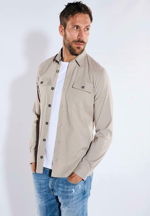 Hellebeige Knopf-bluse mit zwei Vordertaschen und dunklen Knöpfen, getragen über einem weißen T-Shirt, kombiniert mitausgewaschenen blauen Jeans.